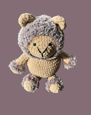 Beiger gehäkelter Amigurumi-Bär mit flauschiger graubrauner Kapuze und grünen Augen vor violettem Hintergrund.