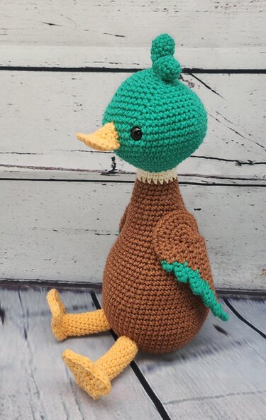 Gehäkelte Amigurumi-Ente mit grünem Kopf, braunem Körper, gelbem Schnabel und gelben Beinen, seitliche Ansicht