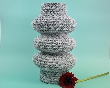 Grau gehäkelte Vase mit gebauchten Ringen und einer roten Schnittblume auf türkisem Untergrund