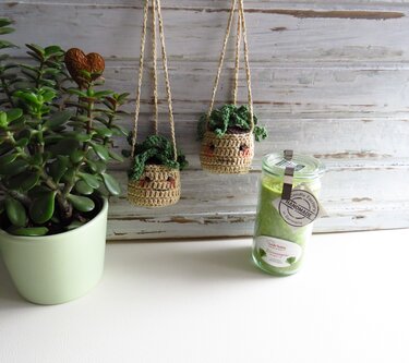 Zwei gehäkelte Amigurumi-Hängetöpfe mit bestickten Gesichtern und kleinen grünen Pflanzen, hängend vor heller Holzvertäfelung; links eine Sukkulente, rechts ein Glas mit Kerze.
