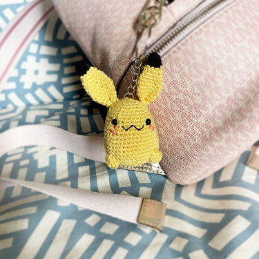 Yellow crocheted amigurumi character keychain with black-tipped ears, wavy embroidered smile, and rosy cheeks attached to a pink bag.