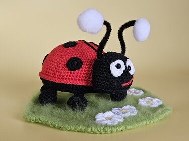 Gehäkelter roter und schwarzer Marienkäfer-Amigurumi mit weißen Pompon-Antennen auf grüner Häkelunterlage mit weißen Blüten