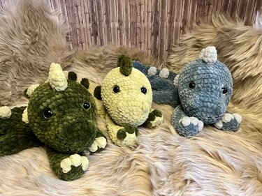 Drei gehäkelte Dino-Amigurumis in Dunkelgrün, Hellgrün und Hellblau auf flauschigem Untergrund