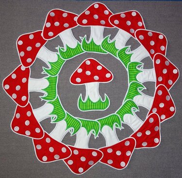 Verspielter Pilzkranz mit roten, weiß gepunkteten Pilzhüten und grünen Blättern auf grauem Stoff