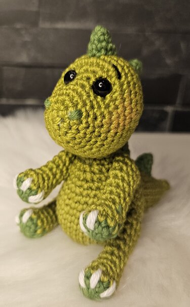 Gehäkelter grüner Amigurumi-Drache mit schwarzen Augen und weißen Krallen, sitzend