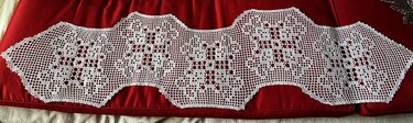 White filet crochet table runner with scalloped wavy edges on a red background.