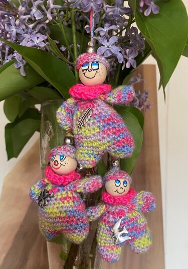Drei gehäkelte Amigurumi-Schlüsselanhänger in Pastellfarben mit Holzköpfen und Metallanhängern vor Fliederzweigen
