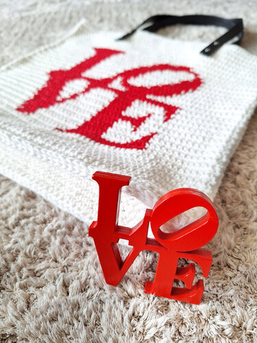Weißer gehäkelter Shopper mit rotem LOVE-Schriftzug und schwarzem Henkel, rotes LOVE-Dekoobjekt im Vordergrund auf Teppich