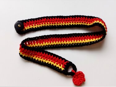 Gehäkelter Riemen in Schwarz, Rot und Gelb mit schwarzem Knopf und kleinem roten Herz auf weißem Hintergrund