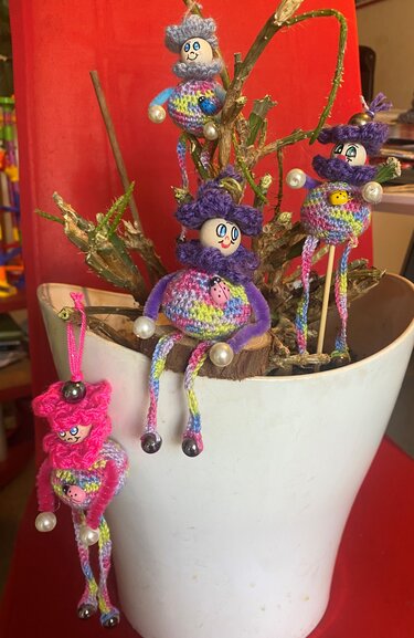 Mehrere kleine gehäkelte Amigurumi-Figuren in Regenbogenfarben mit Holzköpfen und Aufhängeschlaufe, an getrockneten Ästen in einem weißen Blumentopf arrangiert
