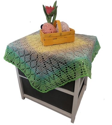 Quadratisches gehäkeltes Deckchen mit Spitzenmuster und gelb-grünem Farbverlauf auf einer kleinen Kommode, dekoriert mit einem Korb und einer Tulpe