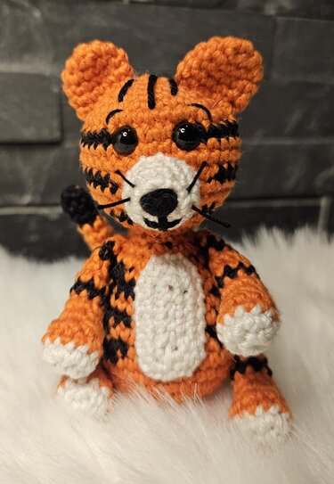 Nahaufnahme eines gehäkelten Amigurumi‑Tigers mit orange‑schwarzen Streifen und weißen Details, sitzend auf weißem flauschigem Untergrund.