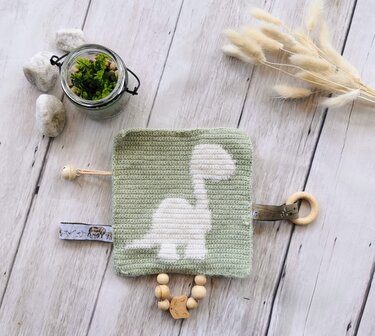 Green crocheted square baby toy with white dinosaur appliqué, wooden teething ring, wooden beads, a small bell, and a patterned fabric tag.