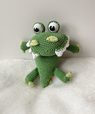 Gehäkeltes grünes Amigurumi‑Monster mit großen Augen und weißen Zähnen auf hellem Stoff