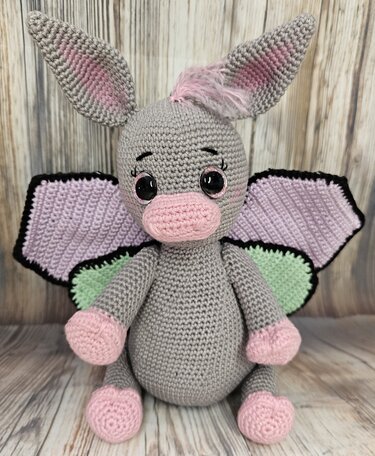Gehäkelter grauer Esel mit rosa Schnauze, Pfoten und Ohren sowie lila-grünen Schmetterlingsflügeln.