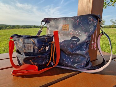 Zwei Umhängetaschen mit floralem und abstraktem orangefarbenem Muster auf einem Holztisch vor einer Grünfläche.