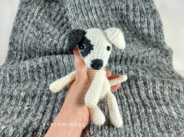 Hand holding small white crochet amigurumi dog with black eye patch and black snout on grey knit blanket