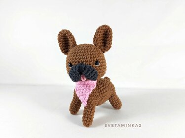 Brown crocheted amigurumi dog with black snout and pink bandana on white background