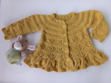 Senfgelbe gestrickte Babyjacke mit Lochmuster, fünf Knöpfen und Rüschensaum, daneben ein kleines Kuscheltier