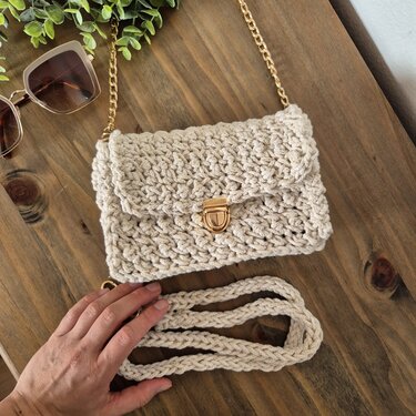 Beigefarbene gehäkelte Clutch mit goldener Kette und Drehverschluss auf Holztisch, daneben gehäkelter Trageriemen und Sonnenbrille.