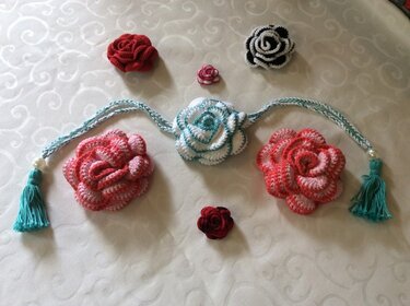Assorted crocheted roses in red, pink, white, teal, and black on patterned fabric, with a braided garland and tassels.