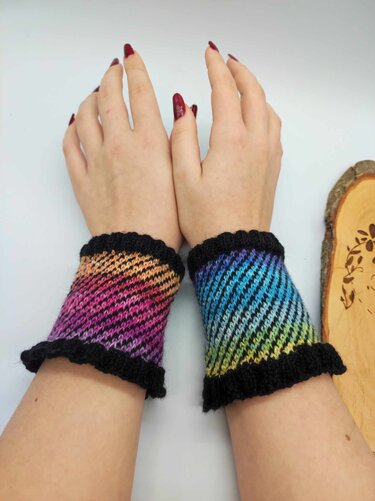 Hände mit bunten, diagonal gestrickten Armstulpen in Regenbogenfarben und schwarzem Rüschenrand