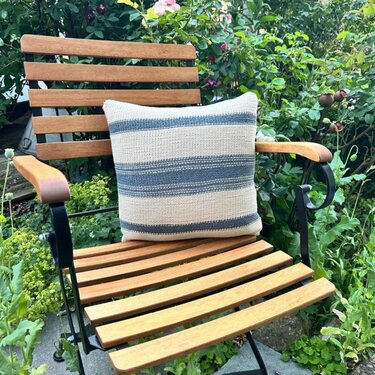 Gestricktes Kissen mit blauen und beigen Streifen auf einem Holz-Gartenstuhl vor grünem Beet