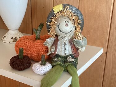 Herbstliche Deko mit gehäkeltem Kürbis-Set in Orange, Braun und Weiß auf einem Regal neben einer Vogelscheuche