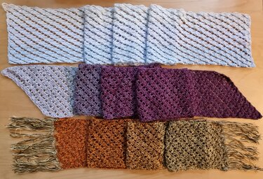 Drei gehäkelte Schals (hellgrau, violett, braun) mit diagonalem Lochmuster auf Holztisch, mittleres Dreieckstuch, unterer Schal mit Fransen