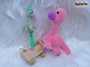 Pastellrosa flauschiger Dino-Amigurumi mit violetter Schnauze und Pfoten neben kleinem grünen gehäkelten Stiftaufsatz auf einem Holzwagen vor weißem Fell
