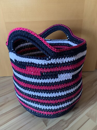 Runder gehäkelter Korb mit horizontalen Streifen in Marineblau, Weiß und Pink und zwei Griffen auf Holzboden