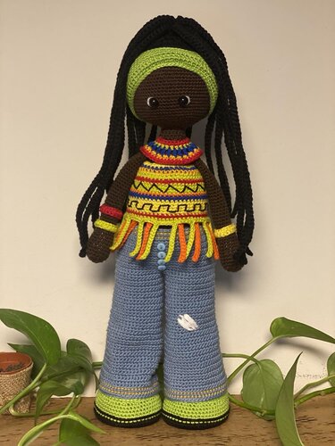 Crocheted amigurumi doll with long black braids, green headband, colorful fringed top and blue flared pants