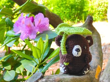Gehäkelter dunkelbrauner Bär auf Ast hält grünes Häkelblatt als Schirm; im Hintergrund grüne Blätter und rosa Rhododendronblüten.