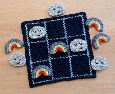 Gehäkeltes Tic-Tac-Toe-Spiel auf hellem Holztisch mit Wolken- und Regenbogen-Spielfiguren, von oben fotografiert.