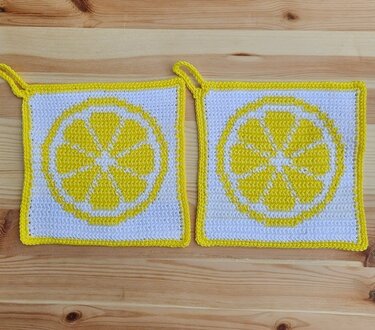 Zwei quadratische gehäkelte Topflappen in Weiß und Gelb mit Zitronenscheibenmotiv auf Holzoberfläche.