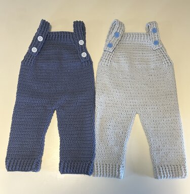 Two crocheted baby overalls, one dark blue with white buttons and one light gray with blue buttons.