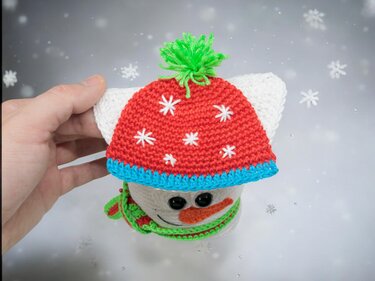 Gehäkelter Schneemannkopf mit roter Mütze und grünem Schal, von einer Hand gehalten, im Hintergrund Schneeflocken