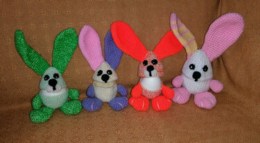 Vier bunte, gehäkelte Hasen-Eierwärmer (Grün, Lila, Orange, Rosa) mit langen Ohren und schwarzen Augen, jeweils ein weißes Ei umschließend.