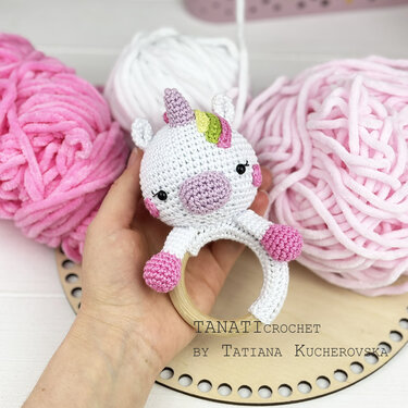 Nahaufnahme eines gehäkelten Einhorn-Greifrings in Weiß und Rosa mit Holzring, gehalten von einer Hand; rosa und weiße Garnknäuel im Hintergrund.
