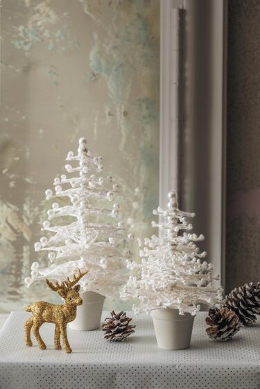 Zwei weiße gehäkelte Mini-Weihnachtsbäume in Töpfen, goldene Hirschfigur und Tannenzapfen auf gepunktetem Tuch.