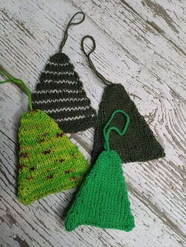 Vier gestrickte dreieckige Anhänger in verschiedenen Grüntönen mit Schlaufen auf hellem Holzuntergrund
