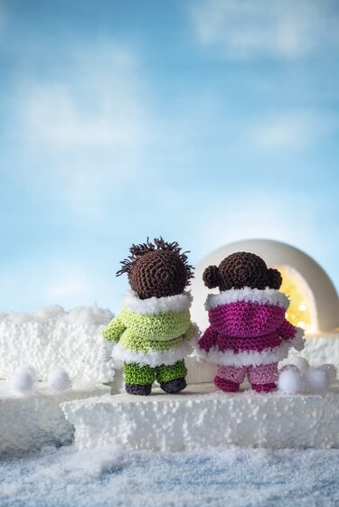 Zwei gehäkelte Amigurumi-Puppen mit grünen und pinken Wintermänteln vor einem beleuchteten Iglu.
