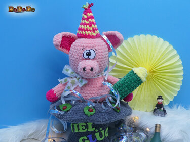 Rosa gehäkeltes Schwein mit gelb‑pinkem Partyhut, grüner gehäkelter Flasche und grauem Sockel mit „VIEL GLÜCK“-Schriftzug.