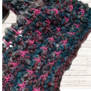 Grob gestrickter Mehrfarbenschal in Grau, Türkis und Beere mit pinken Kreuzstichen auf hellem Holz