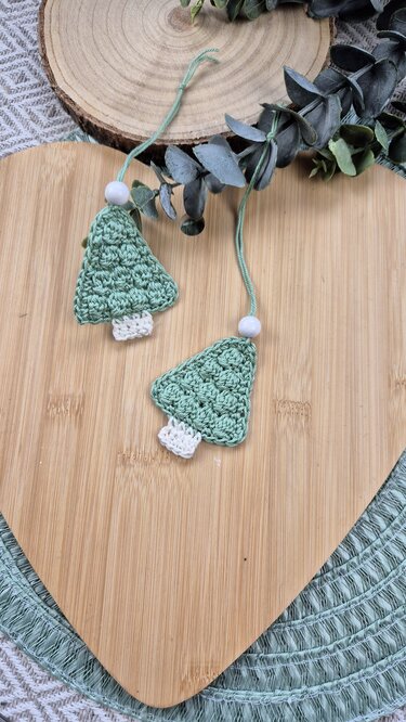 Zwei gehäkelte hellgrüne Tannenbaum-Anhänger mit weißem Stamm und Holzperlen auf Holzbrett.
