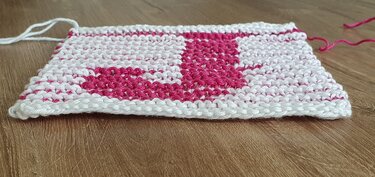 Gestricktes quadratisches Tuch mit pinkem Buchstaben A auf weißem Grund im Kraus‑rechts‑Muster auf Holzuntergrund.