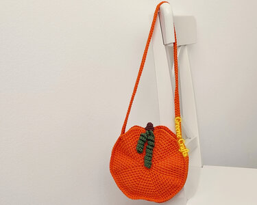 Orange crocheted pumpkin-shaped shoulder bag with green stem and braided strap.