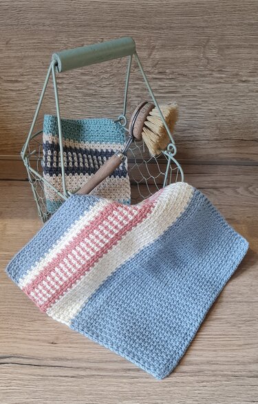 Gehäkeltes Spültuch mit blauen, rosa und weißen Streifen auf Holz; im Hintergrund Drahtkorb mit Tuch und Spülbürste.