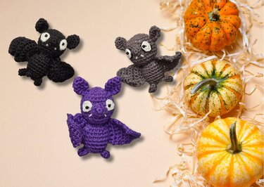 Drei gehäkelte Amigurumi-Fledermäuse (schwarz, grau, lila) neben kleinen Kürbissen