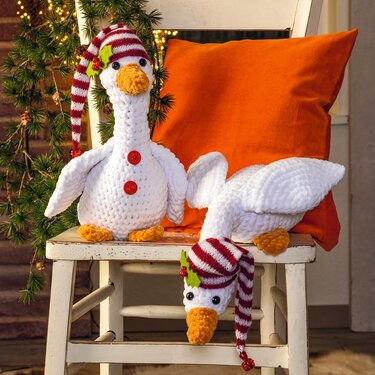 Zwei gehäkelte weiße Gänse mit gestreiften Weihnachtsmützen, orangen Schnäbeln und roten Knöpfen auf einem Stuhl neben Tannengrün